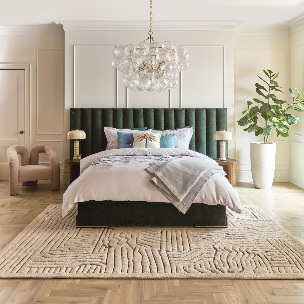 A big bedroom design with a green upholstered bed, wall panelling, statement chandelier and a plush rug.