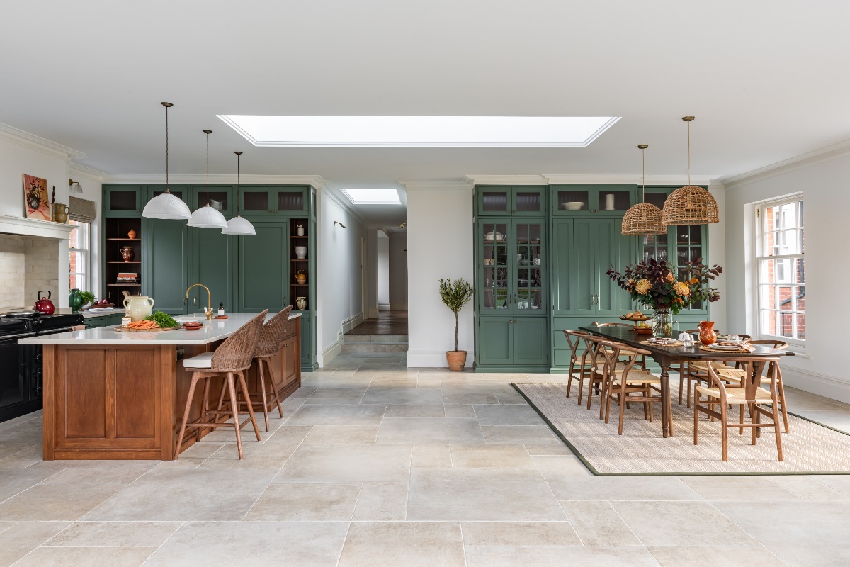 Large open-plan space with an L-shaped kitchen, drinks area and dining zone with stone flooring, crisp white walls and glazing.