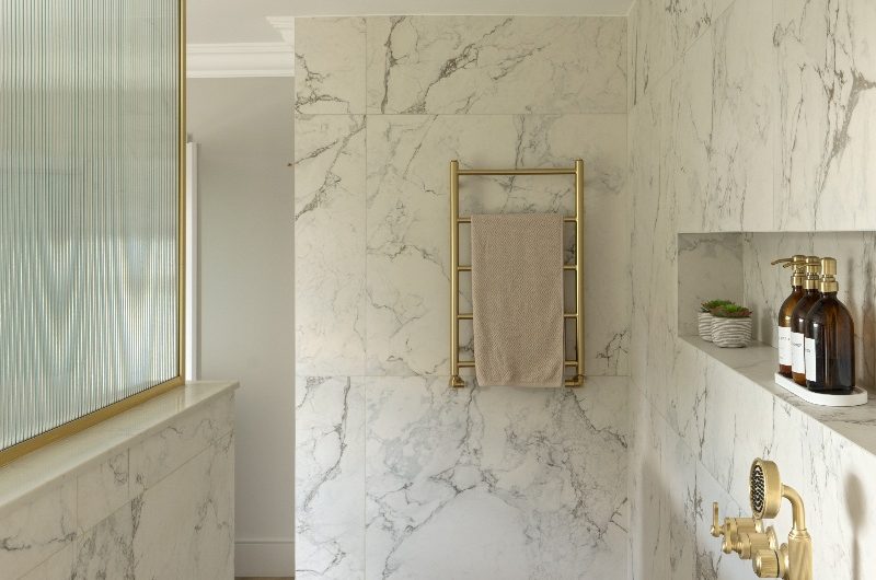 An open shower space with gold brassware, storage niche, towel holder, marble surfaces and a pony wall.
