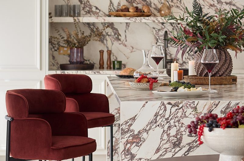 A luxury kitchen design with an island decked out in marble-effect surfaces, a matching splashback with open shelf, white cabinetry, and red velvet bar stools.
