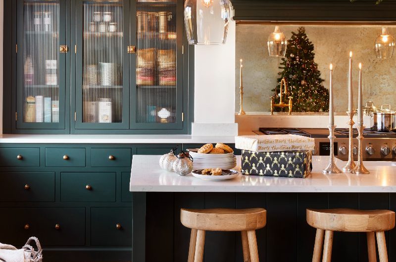 A dark green Shaker kitchen with a glazed fluted glass cupboard, a range cooker zone with mirrored splashback, and a central island with decor.