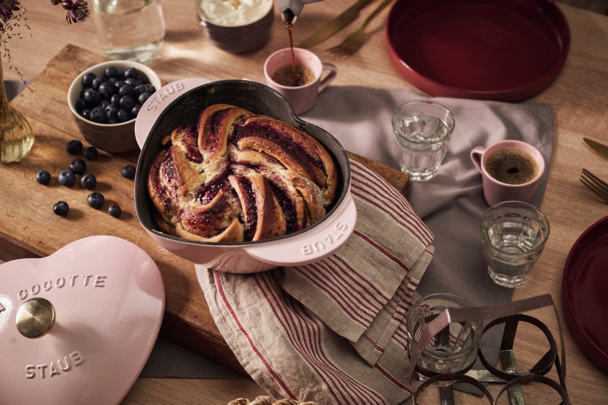 A Christmas tablescape with a heart shaped cocotte, glassware, round burgundy plates, and a wood serving tray.