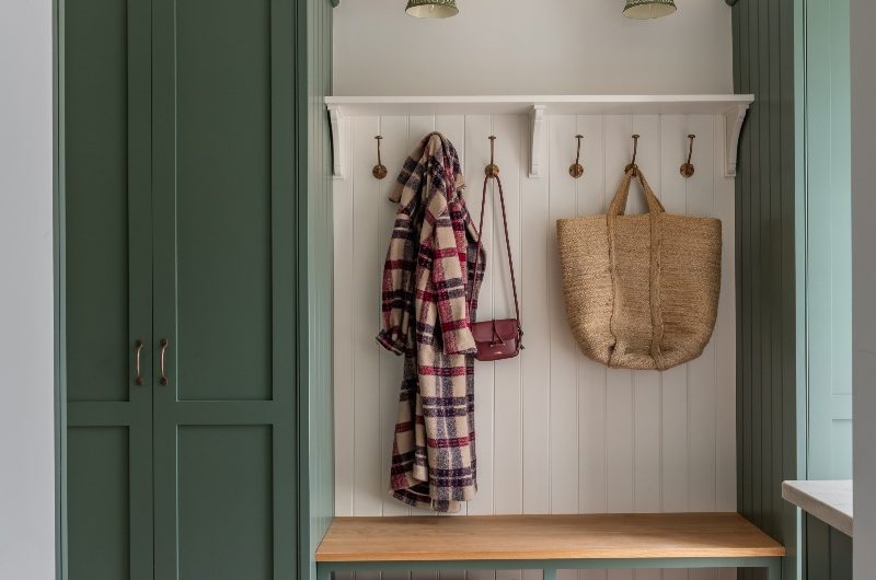 A bootroom with green cabinetry, a bench with a wood top, wall panelling, terracotta flooring, and brass detailing.