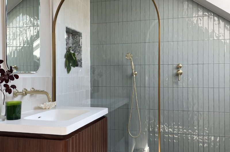A bathroom design with a shower zone, marble floor tiles, glossy white and green metro wall tiles, a fluted wood vanity, and a gold shower enclosure.
