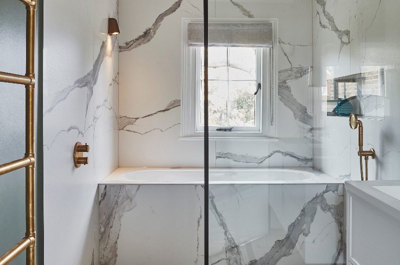 A modern wetroom drenched in veiny wall and floor tiles, with a fitted tub, shower zone, framed by a matt black panel, and a white vanity.