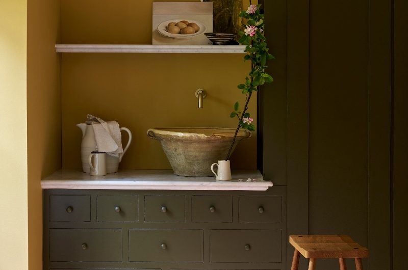 A country kitchen with green cabinetry, a high wood stool, yellow paint on the walls, and a rustic basin.