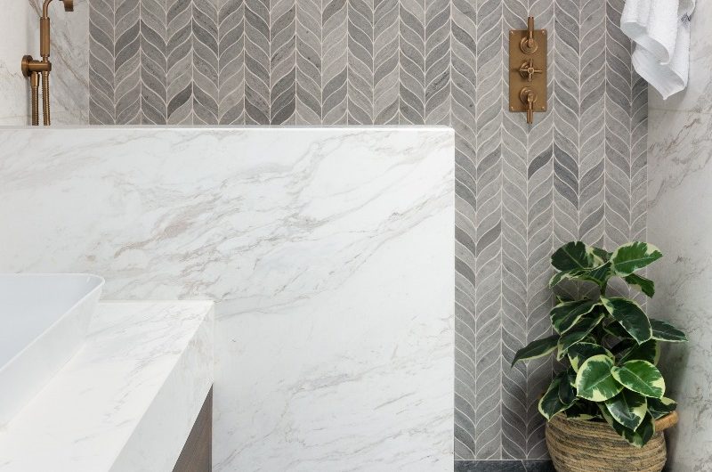 A bathroom scheme with layered materials, such as feather-pattern tiles with cool blue-grey tone, brass fixtures, and timber detailing.