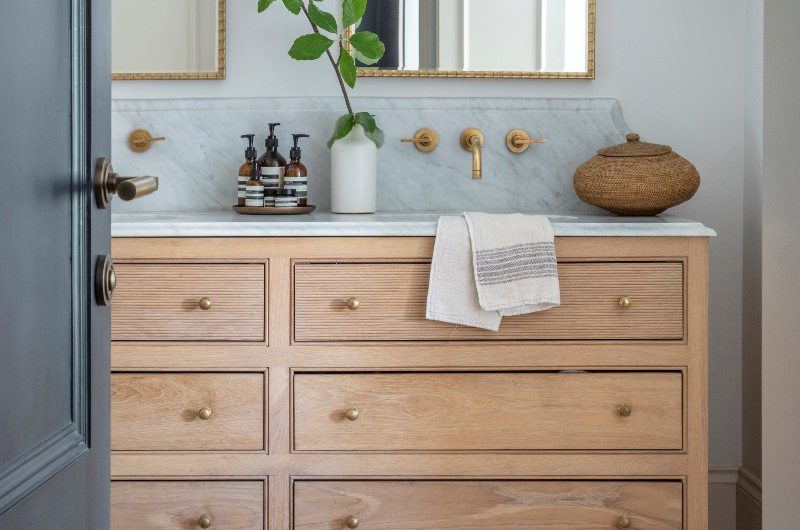 A calm bathroom design with white wall paint, a double light wood vanity unit with marbled worktops and splashback and gold taps, two brass framed mirrors, wall sconces, and terracotta flooring with a durable rug.