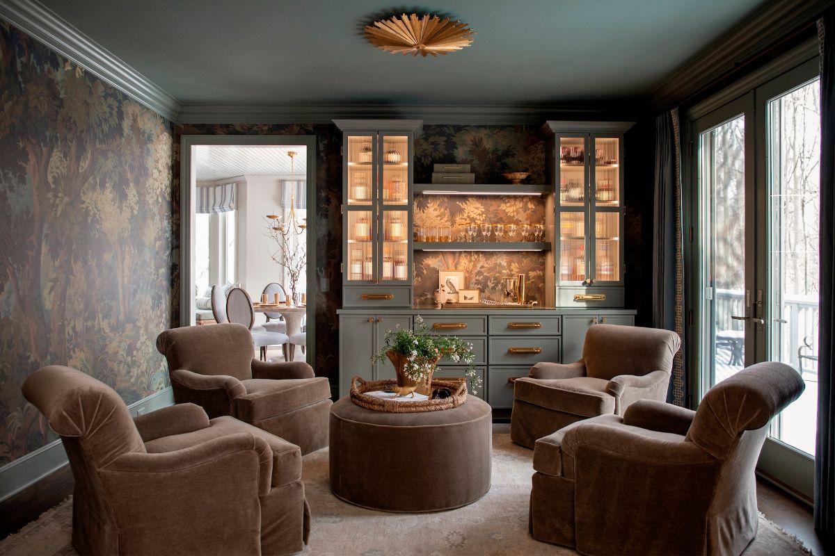 An entertaining zone with dark green paint on the ceiling, patterned wallpaper, a drinks cupboard with LED lighting, and plush, dark brown armchairs and pouffe in the centre.