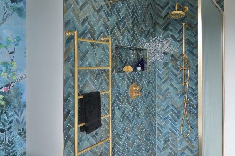 A blue wetroom with herringbone wall tiles, gold shower system, radiator and panel, and marbled flooring.
