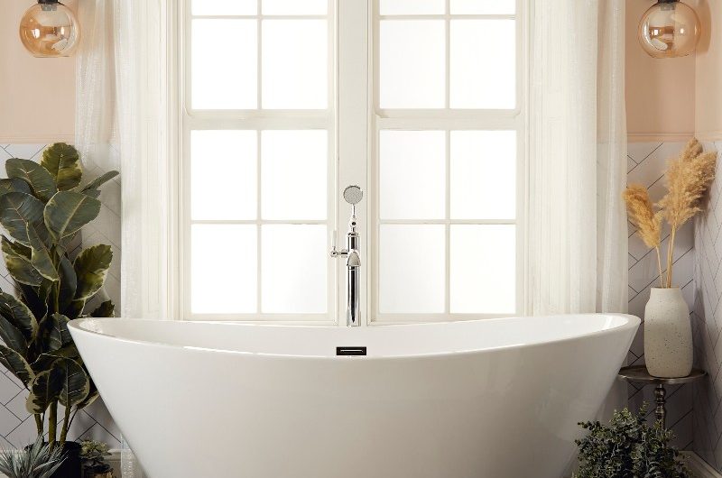 A calm bathroom scheme with a double-ended bath, wood flooring, sheer curtains, and house plants.