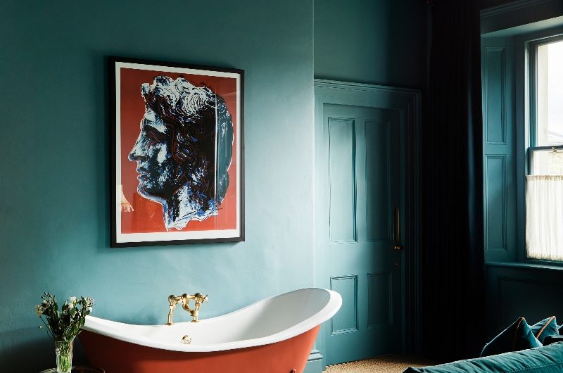 A teal bathroom design with a dark orange bathtub, wall art above it and a velvet sofa.