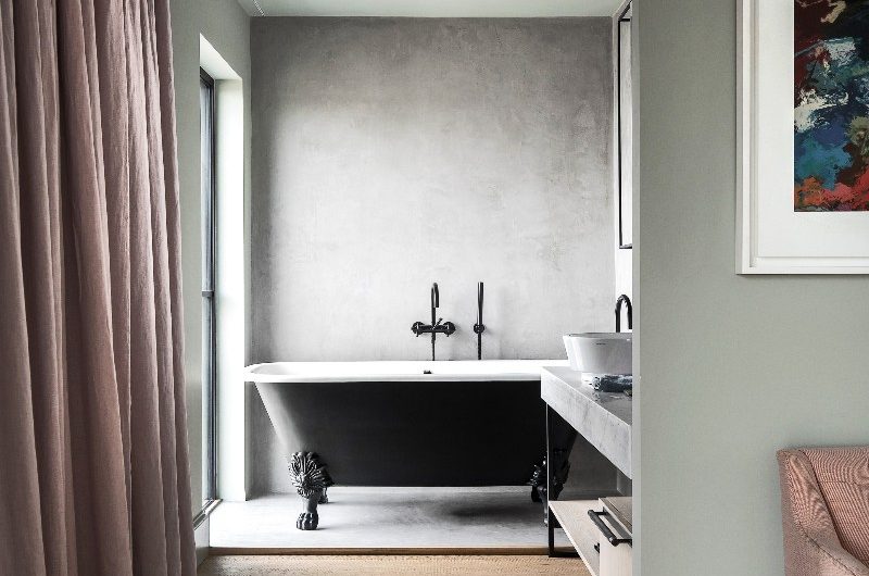 A modern scheme with wood flooring leading to a bathing space with concrete-effect surfaces, a freestanding tub painted in black with bronze brassware.