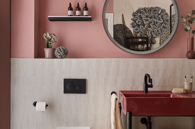 A modern scheme with pink paint on the upper half of the wall, white tiles on the bottom half of the wall, with a glossy red basin on a black steel frame, with a circular mirror above and wall-mounted toilet next to it.