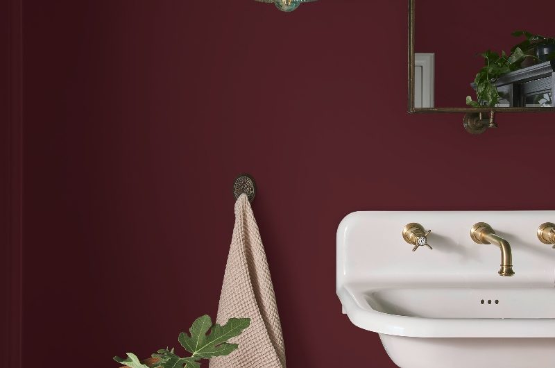 A burgundy bathroom with a wall-mounted basin, brass wall light and mirror, and a wood chair with decor.
