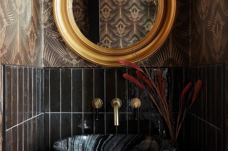An Art Deco cloakroom with dark metro tiles, patterned wallpaper, a gold mirror, a stone basin and gold brassware.
