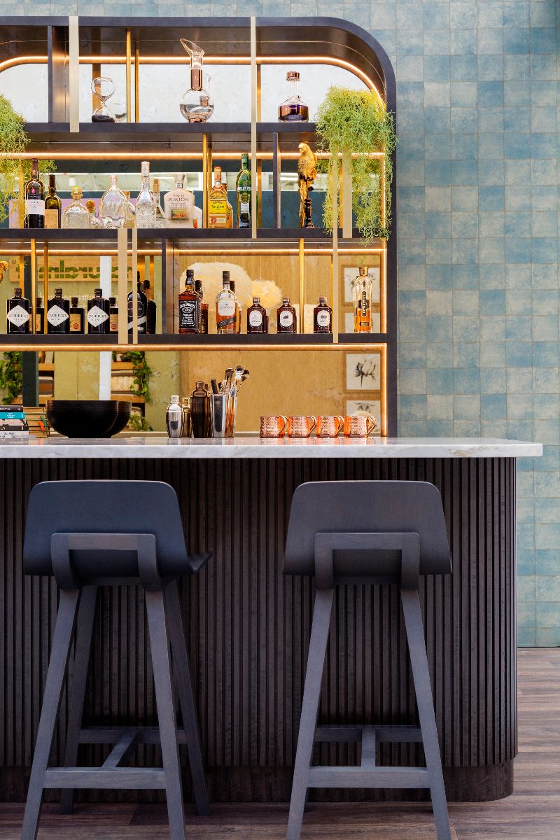 A luxurious home bar design with pale blue zellige tiles, a fluted wood island with marble worktop and bar stools, and a bottle display area with LED lighting and mirrored splashback.