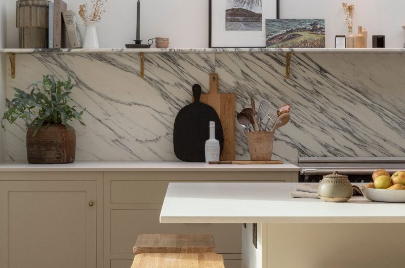 A modern kitchen design with cream cabinetry, statement marble worktop and splashback, and open shelving.
