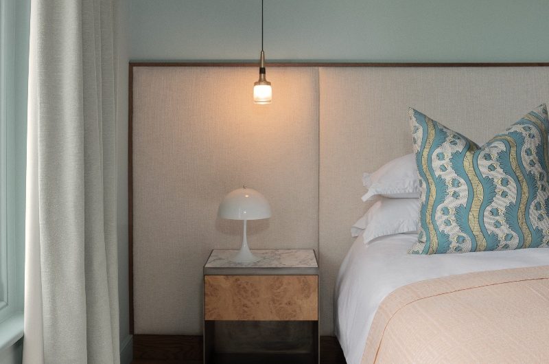 A calm bedroom design with an extra-large headboard, a minimalist bedside table, white bed linen, a blue patterned cushion, a peach throw, and a neutral rug underneath.