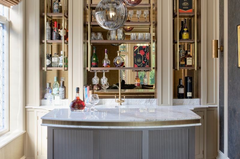 A home bar with a statement pendant, mirrored splashback, an island with fluted base and marbled worktop, and bottles on display in the background.