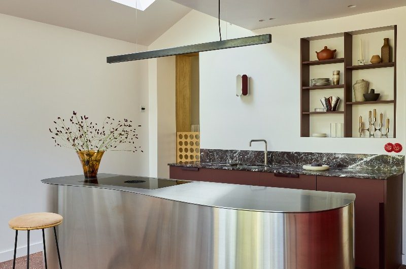 A kitchen design with a restaurant feel, a stainless steel island, a burgundy cabinetry run behind it, and red terrazzo flooring.