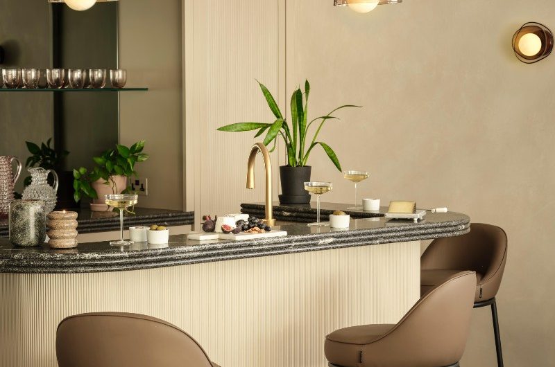 A home bar with statement lighting, dark worktops, a fluted island, and leather bar stools.