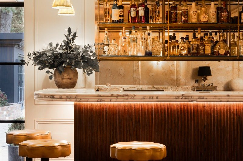 A bespoke home bar with layered lighting, a mirrored splashback, a fluted oak island with statement marble and restaurant-inspired stools.