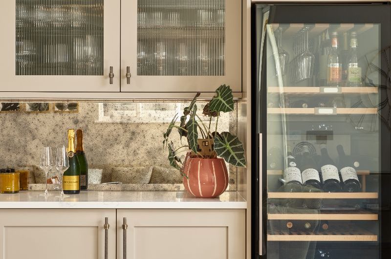 A kitchen design with cream Shaker cabinets, brass handles, a tall wine fridge, aged mirror splashback, and fluted glass wall cabinets.