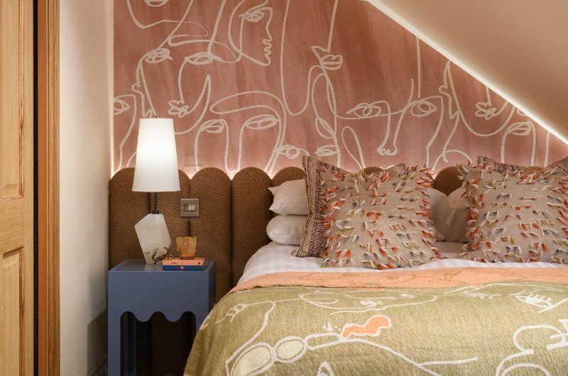 A calm but colourful bedroom with a mix of patterns, from the pale terracotta abstract wallpaper to the green throw, with a scalloped headboard and quirky blue side table.