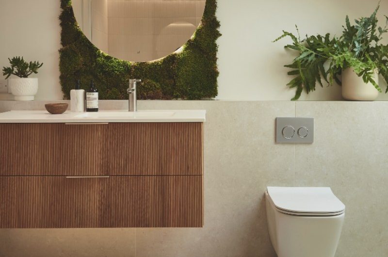 A calm bathroom scheme with nature-inspired wall and floor coverings, a living plant installation, a circular, illuminated mirror, and fluted wood vanity.
