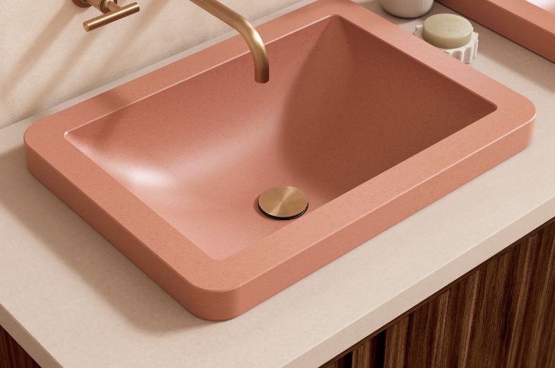 A modern bathroom with a wood vanity with cream countertop and coral inset basin, with gold detailing and matching taps.