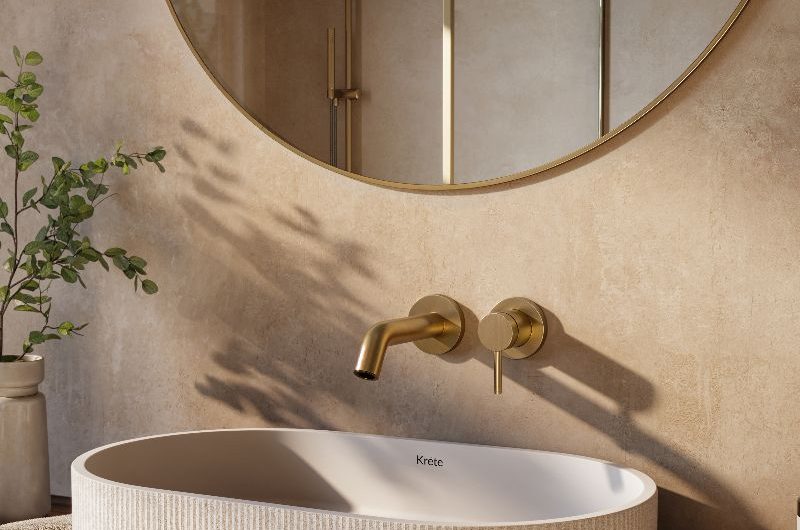 A neutral bathroom design with a tactile basin, brass tao, plaster walls and a brass circular mirror above it.