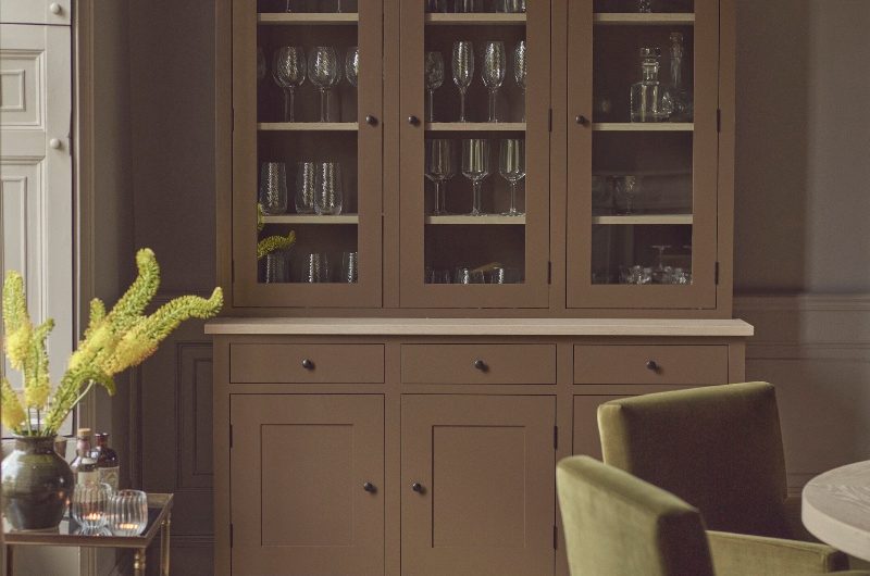 A dining room with a brown glassware dresser, green velvet chairs, a wood table and grainy wood flooring.