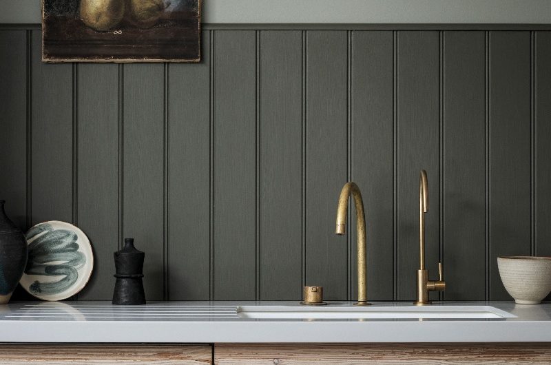 A moody kitchen design with light wood cabinets, dark green panelled walls, brass taps, and rustic decor.