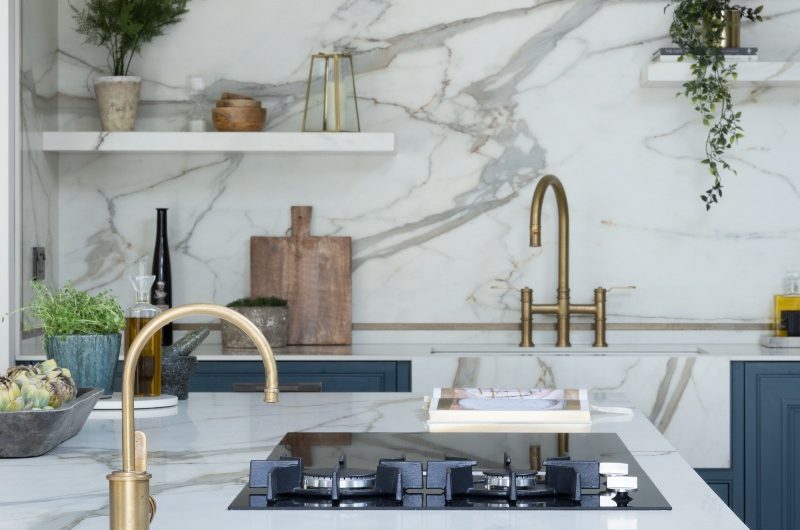 View of the sink area from the island with a wood breakfast bar, a tiny sink and brass tap, a gas and induction hob, and matching porcelain surfaces.