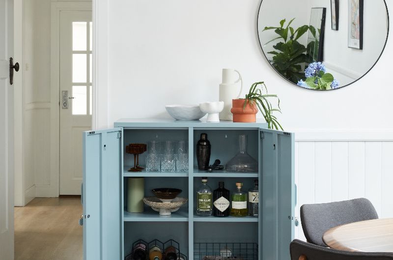 A bright dining room with a compact locker-style cupboard in pale blue storing glasses and bottles.