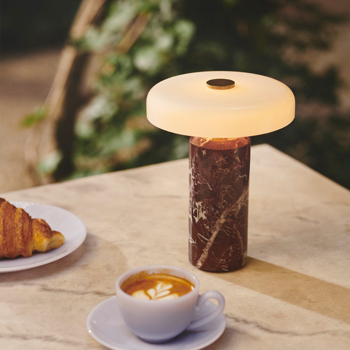 A stone table with croissant and cappuccino and a marble and opal glass table lamp.
