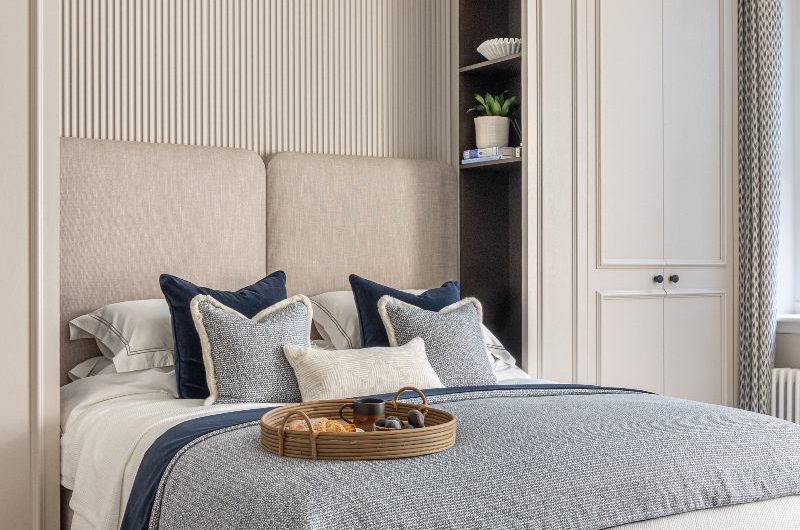 A bright, off-white scheme with panelled wardrobes, fluted paneling, and a statement bed frame in neutral upholstery fabric, decorated with white and blue bedding.