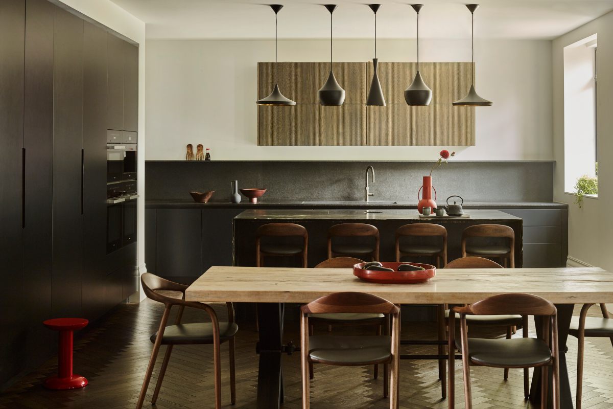 A dark kitchen design with handleless cabinetry, a central island, and a dining zone with a wood table and mid-century style chairs.