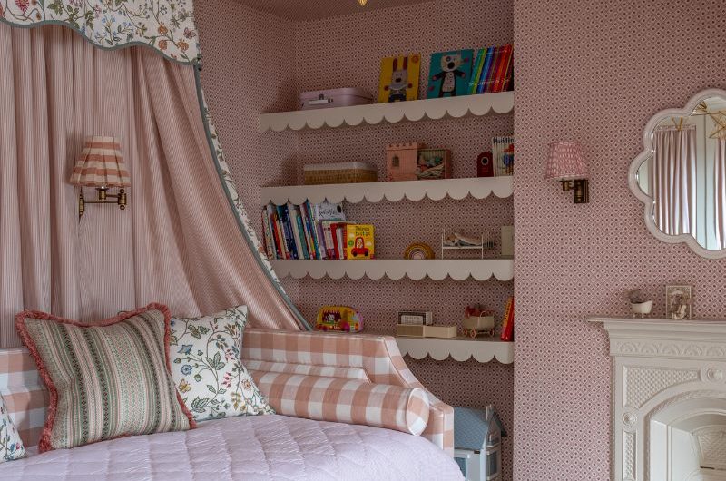 A girl's bedroom with gingham canopy bed with pink wallpaper, white scalloped shelved with kids toys and books, and a playful rug.