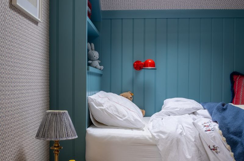 A boys bedroom with a panelled bed, white linen, panelled wallpaper, and a wood stool with a table lamp on top of it.