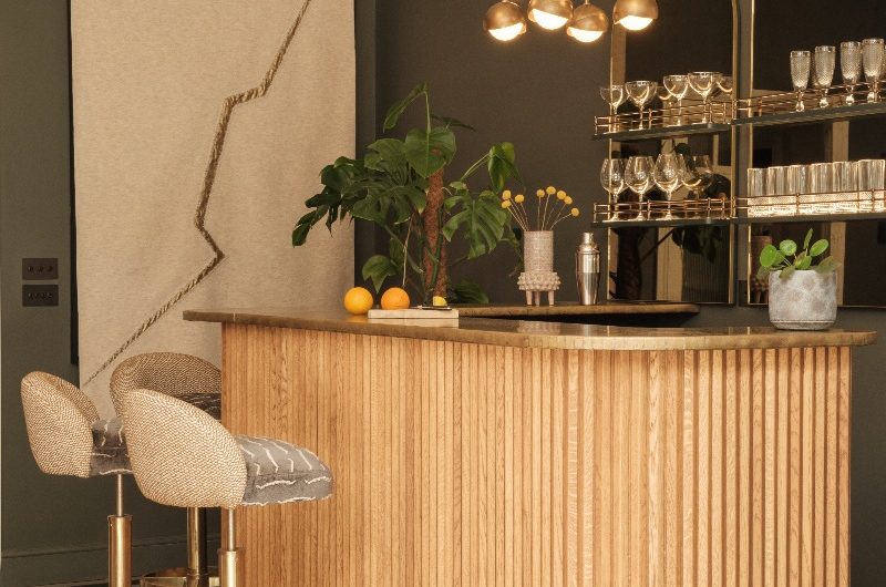 A glamorous home bar with fluted wood island, brass tabletop, stylish bar stools, and glassware display.