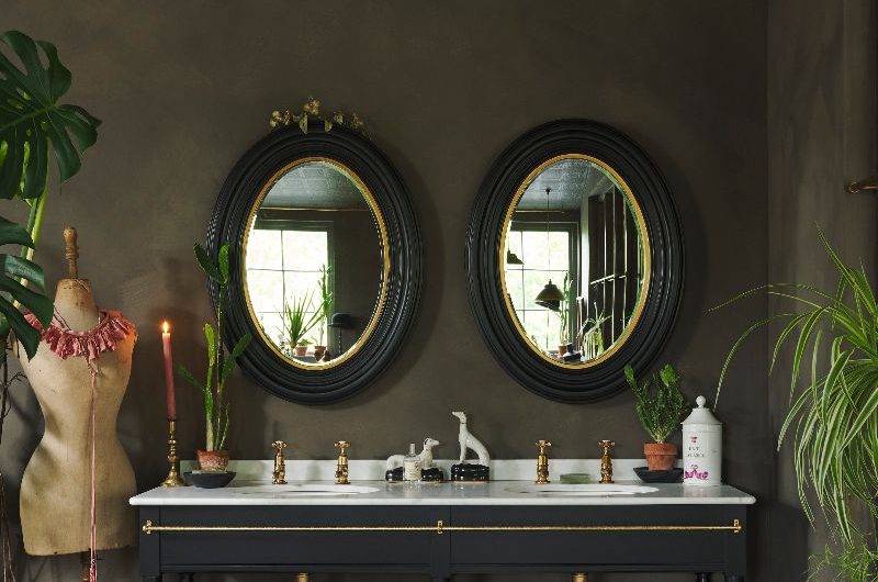 An eclectic bathroom design with dark green wall paint, a black double vanity unit with inset basins and statement twin mirrors above it.