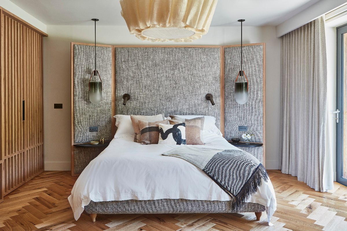 A statement scheme with a bespoke headboard with heavily textured fabric, wood herringbone flooring, bold pendant lights, and white linen.
