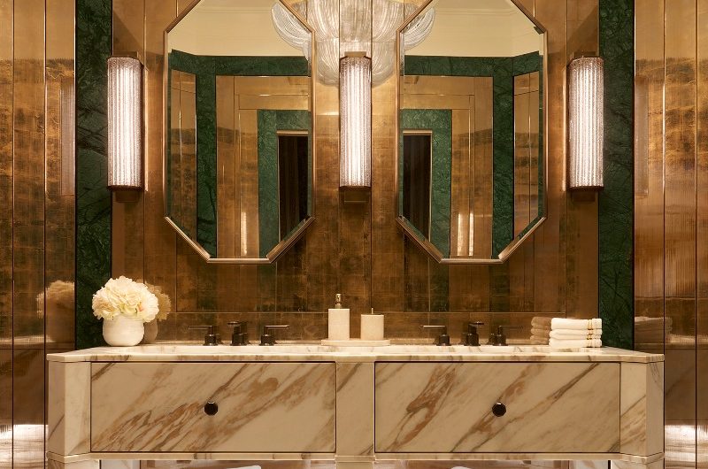 A luxurious bathroom vanity area made from marble with drawers, open storage, two brass mirrors and metallic wall cladding.