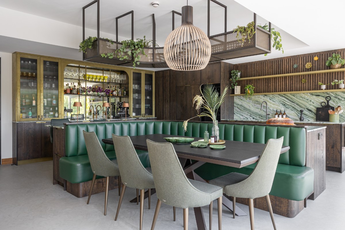 A modern kitchen with a multi-functional island hub which has a green marble worktops, a green leather banquette with dining table and chairs, a hob and hanging display area for house plants above it.
