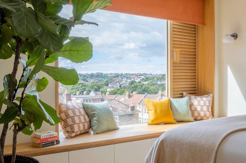 A window nook with a built-in bench with white handleless drawers and w wood top, with an orange roller blind and colourful cushions.