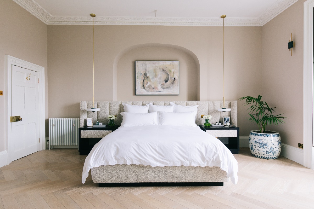 A large neutral bedroom with a centrally placed bed in front of a wall recess, with an oversized boucle headboard, white linen, twin bedside tables and pendants, and wood flooring.