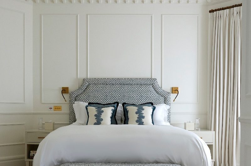 A white, sophisticated design with elegant wall panelling, a central bed upholstered in a blue fabric with a matching ottoman and neutral bedside storage.