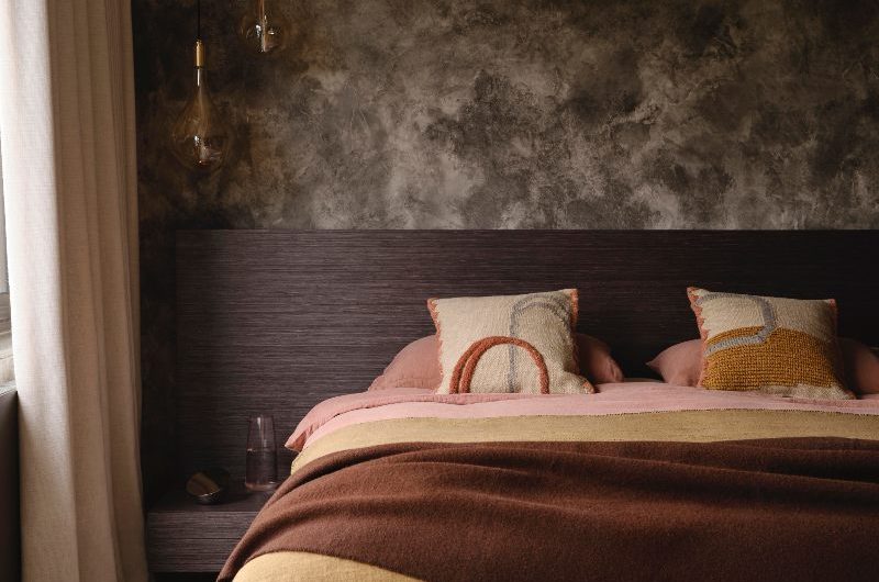 A moody sleep space with a wood headboard and built-in bedside table, dark wallpaper, warm toned soft furnishings.